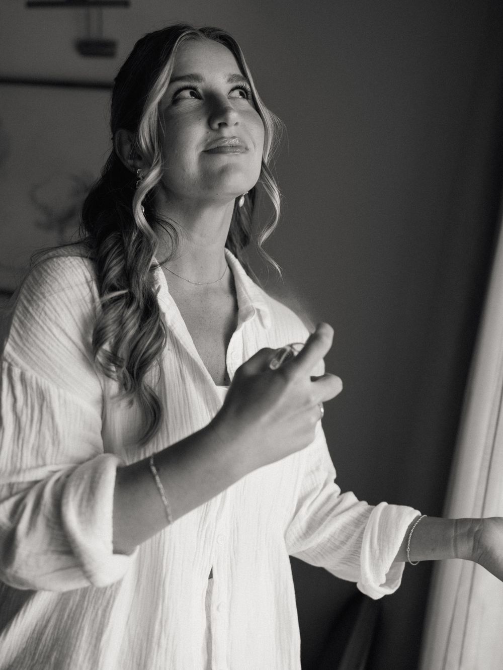 A black and white portrait of a bride in a white blouse gazing softly near a window.