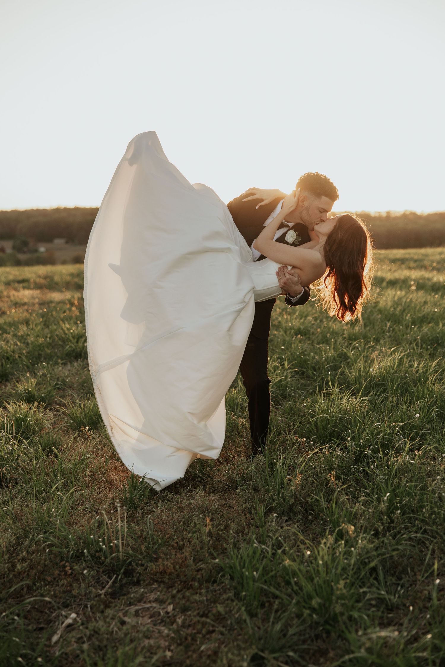 Couple in wedding attire share romantic dip kiss in golden sunlit field at sunset.