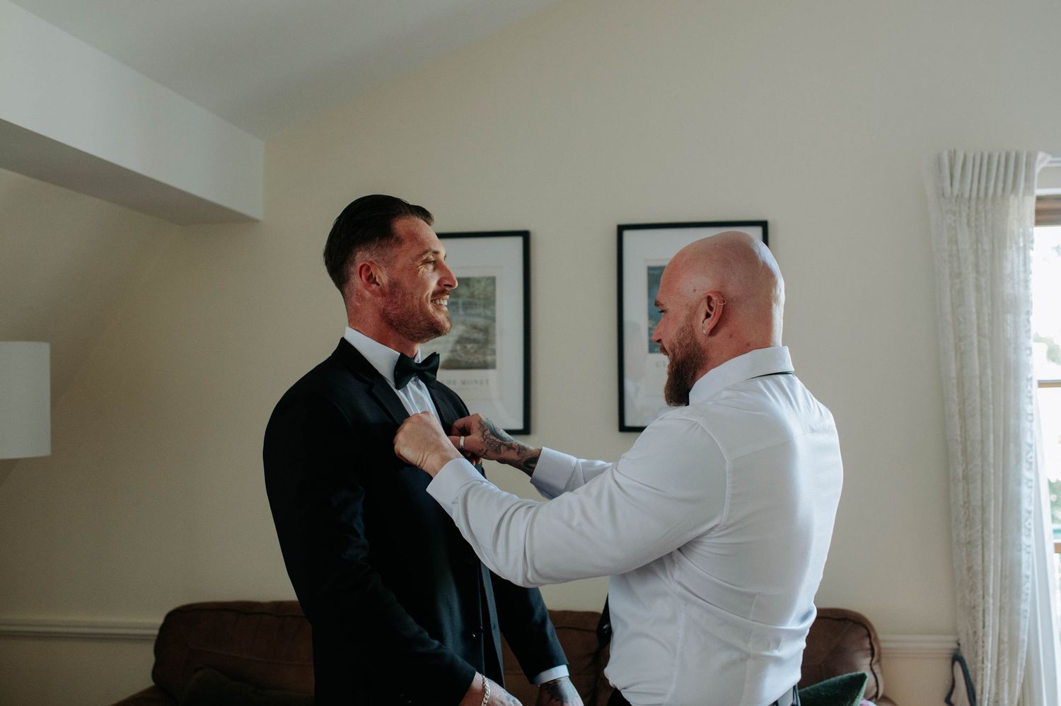 Man adjusting another man's black tie while getting ready for formal event in bright room.