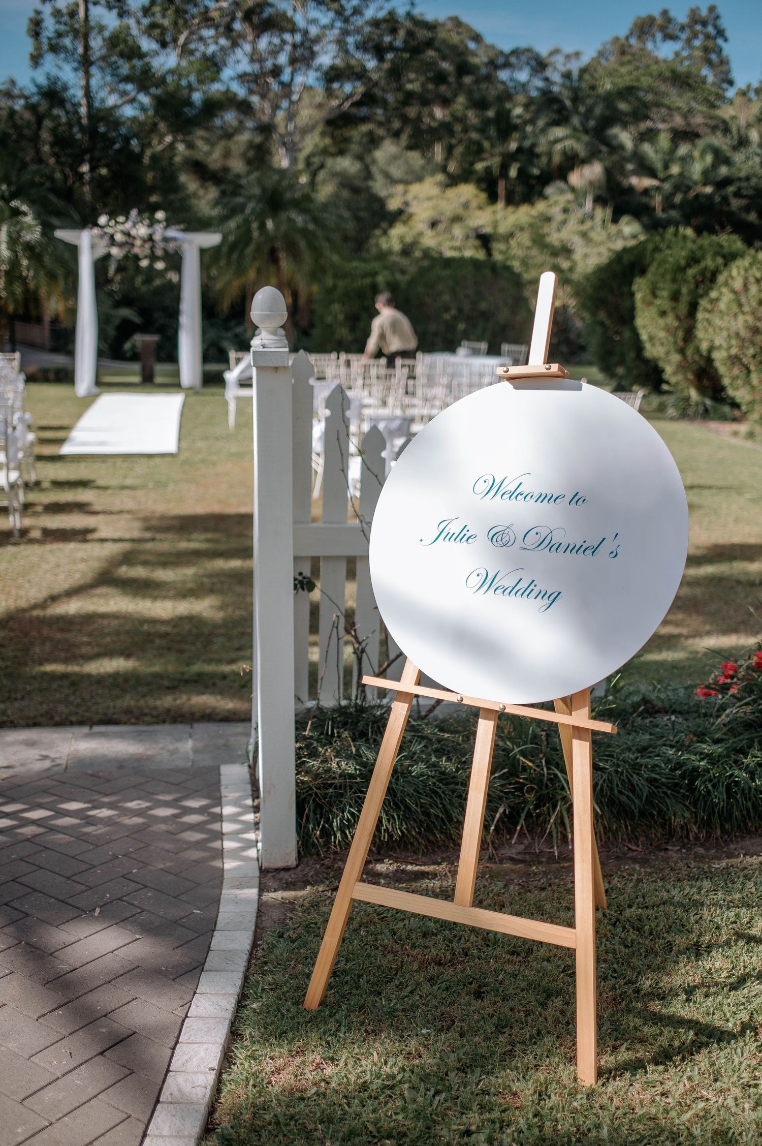 A series of white wedding welcome signs on wooden easels displayed along a garden pathway at an outdoor venue.