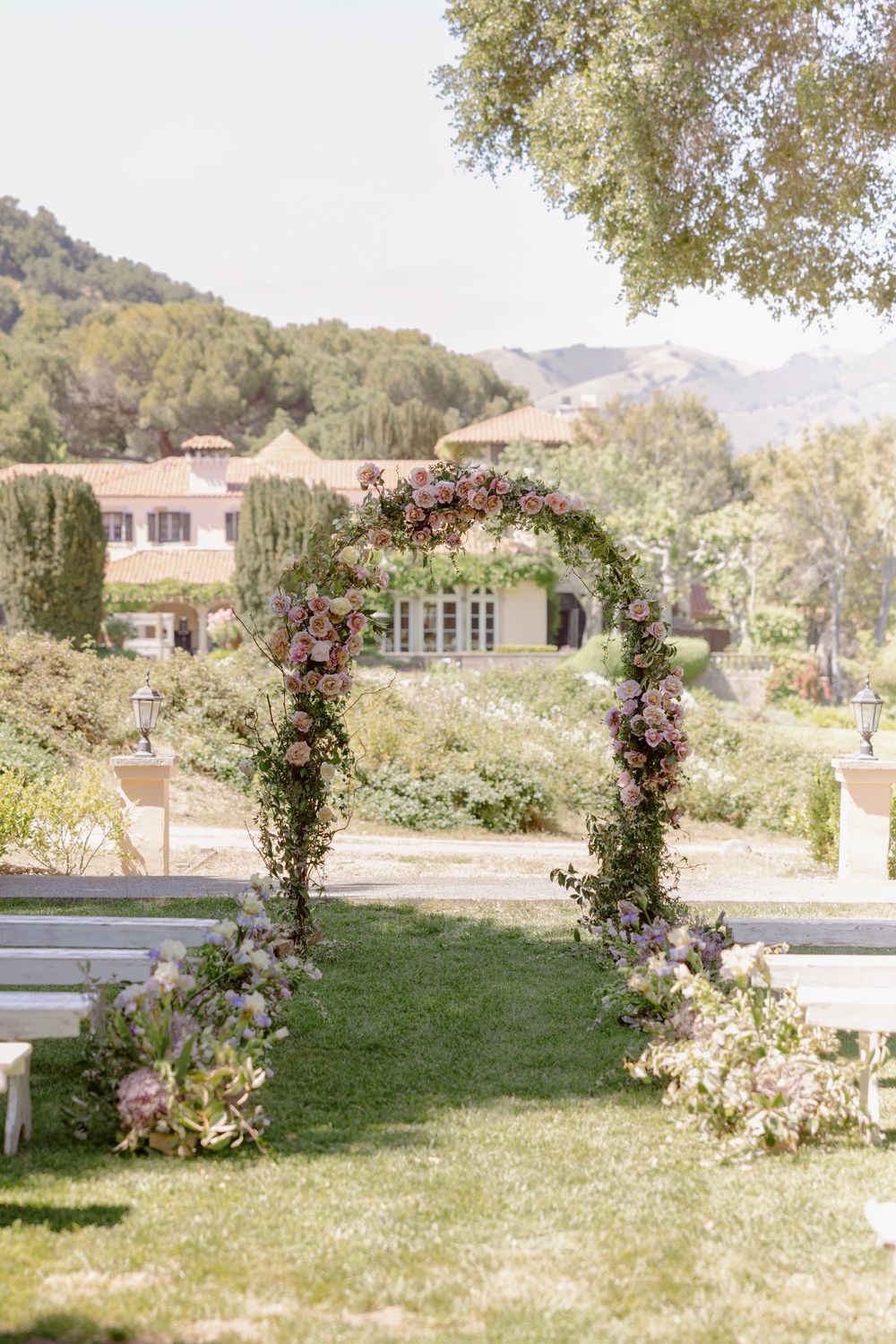 A romantic floral wedding arch with pink and purple blooms stands on a manicured lawn with mountain views in the background.
