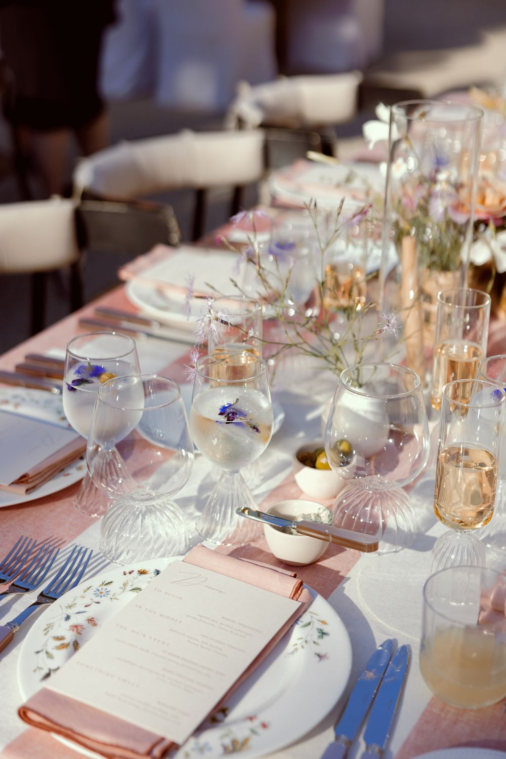 Elegant wedding reception table setting with floral china plates, crystal glasses and delicate pink accents at sunset.