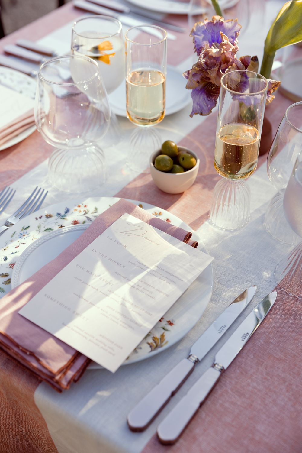 Elegant table setting with pink linens, champagne glasses, menu cards, olives and purple floral arrangements.