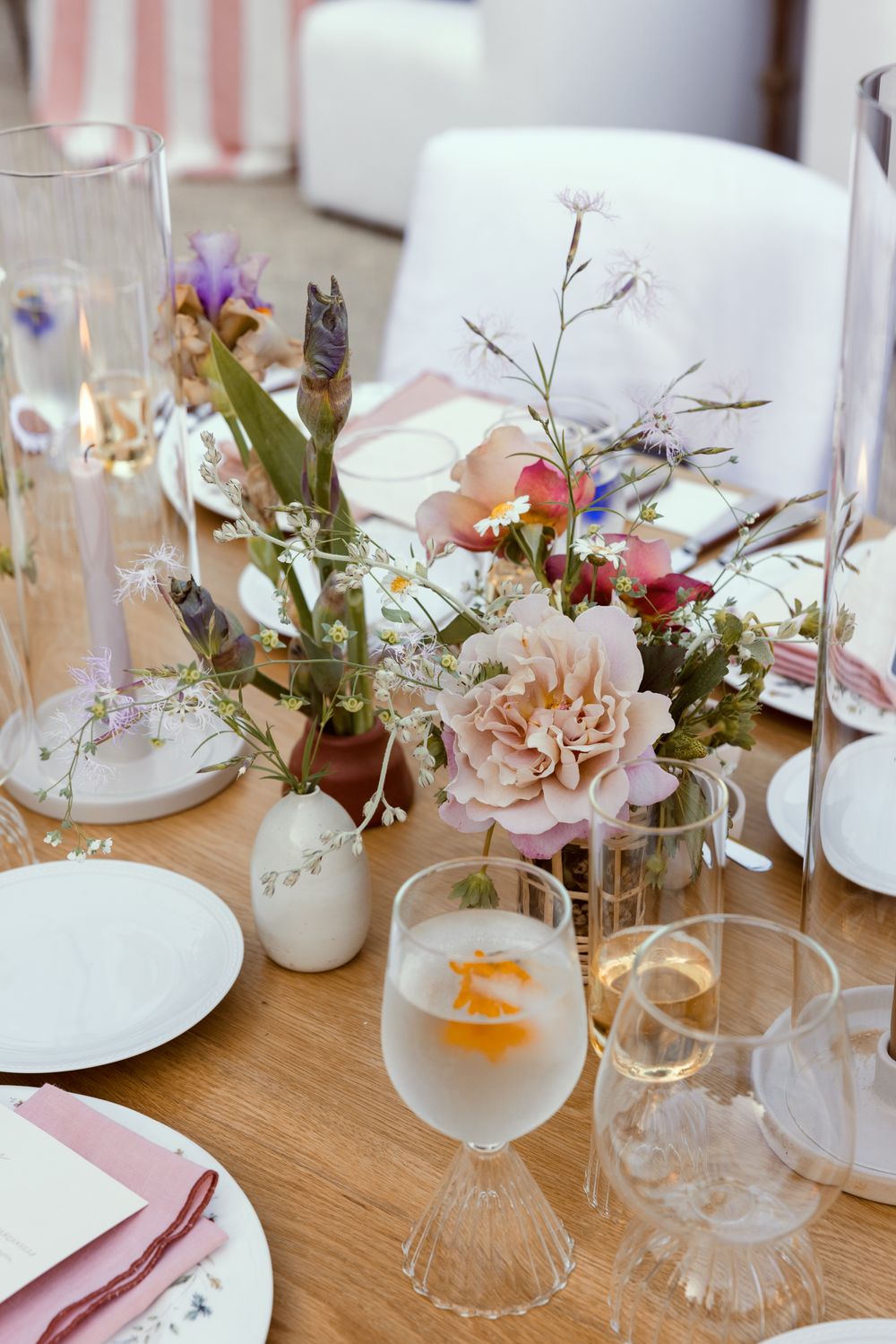 Elegant table setting with soft pink floral centerpiece, white plates, and crystal glasses on wooden surface.