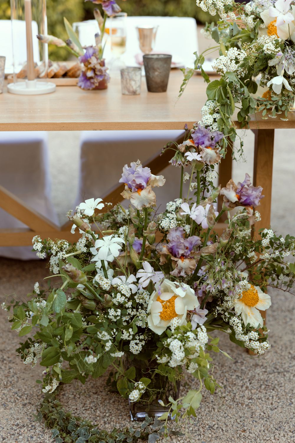Delicate floral arrangement with white daisies, purple iris and baby's breath on a rustic outdoor dining table setting.