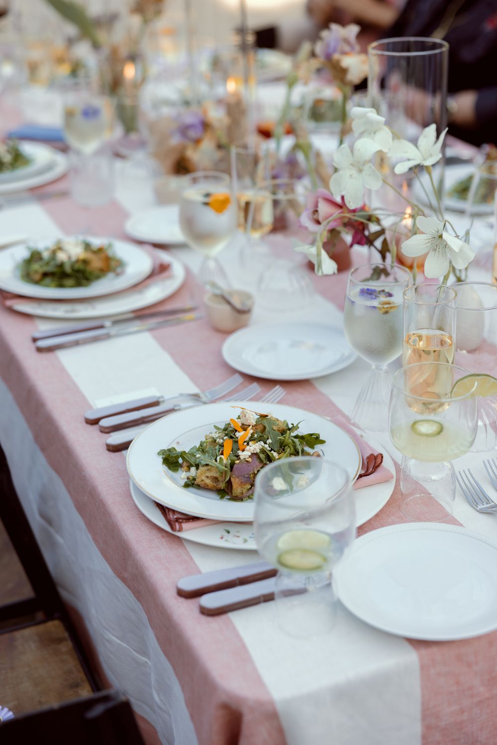 A formal dining table setting with pink tablecloth, white plates, elegant glassware and delicate floral centerpieces.