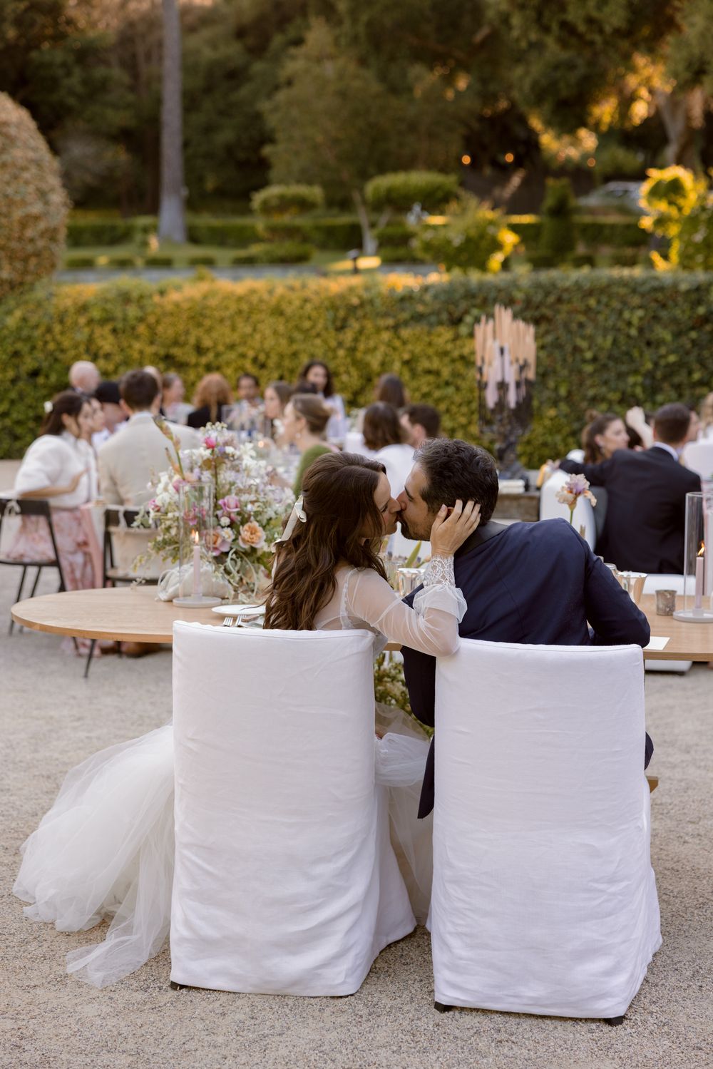 Newlyweds share an intimate moment while seated in white covered chairs at an outdoor garden reception with guests behind them.