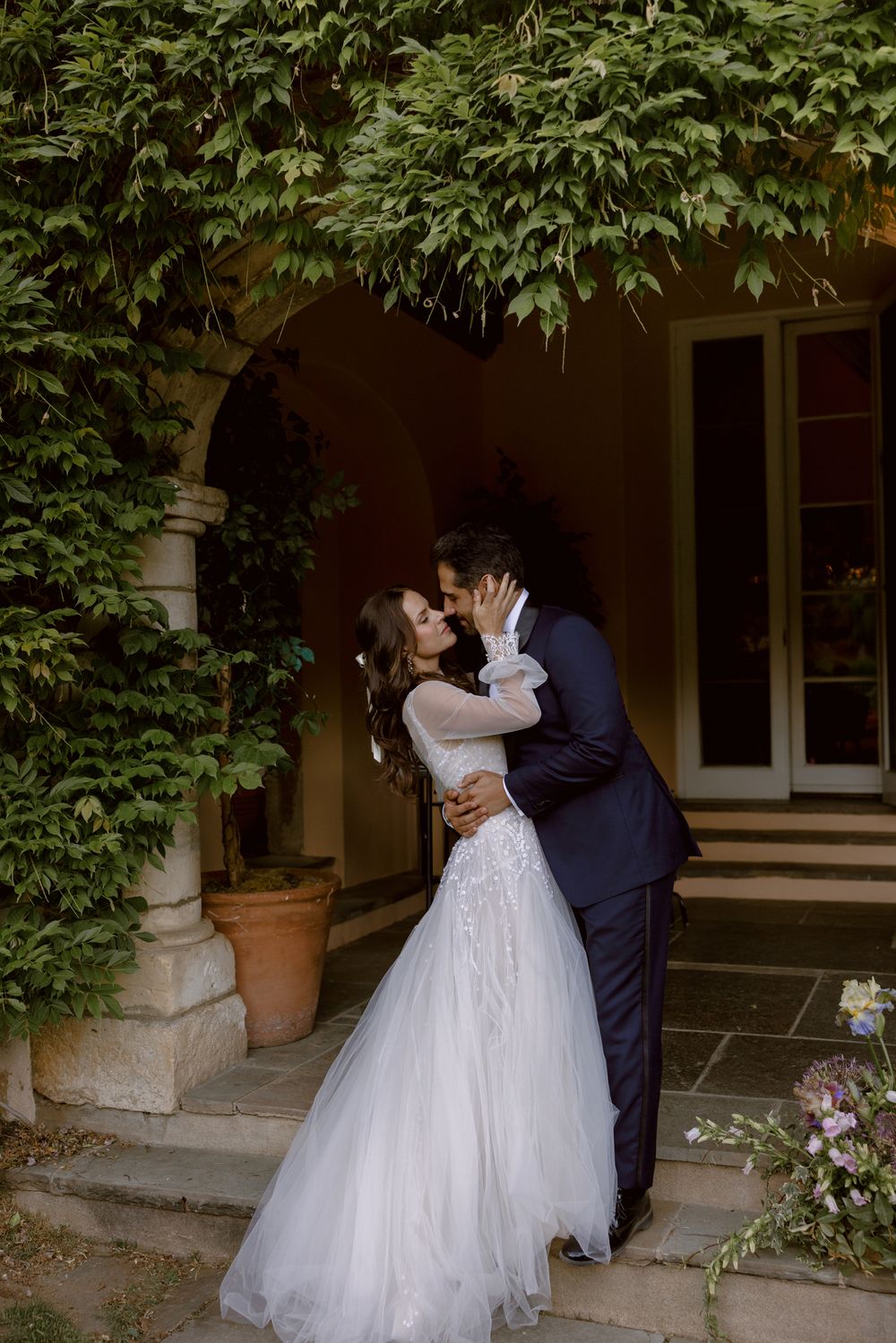 A romantic couple share a kiss under a stone archway adorned with lush green vines and flowers.