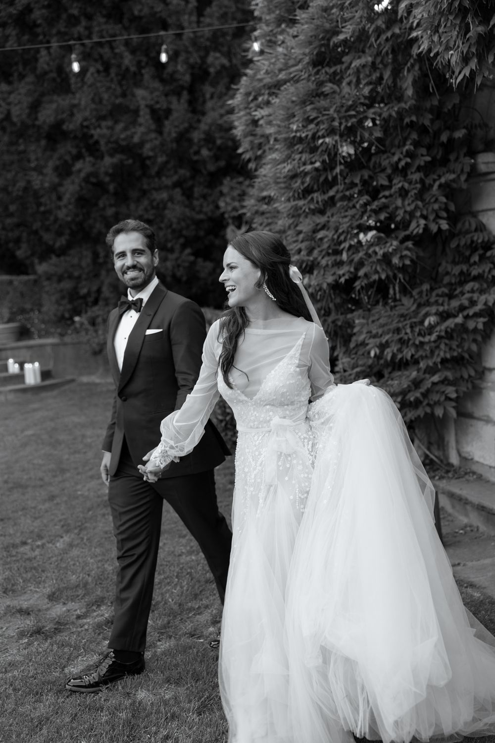 A black and white outdoor wedding photo shows a couple walking together on a lawn with string lights overhead.