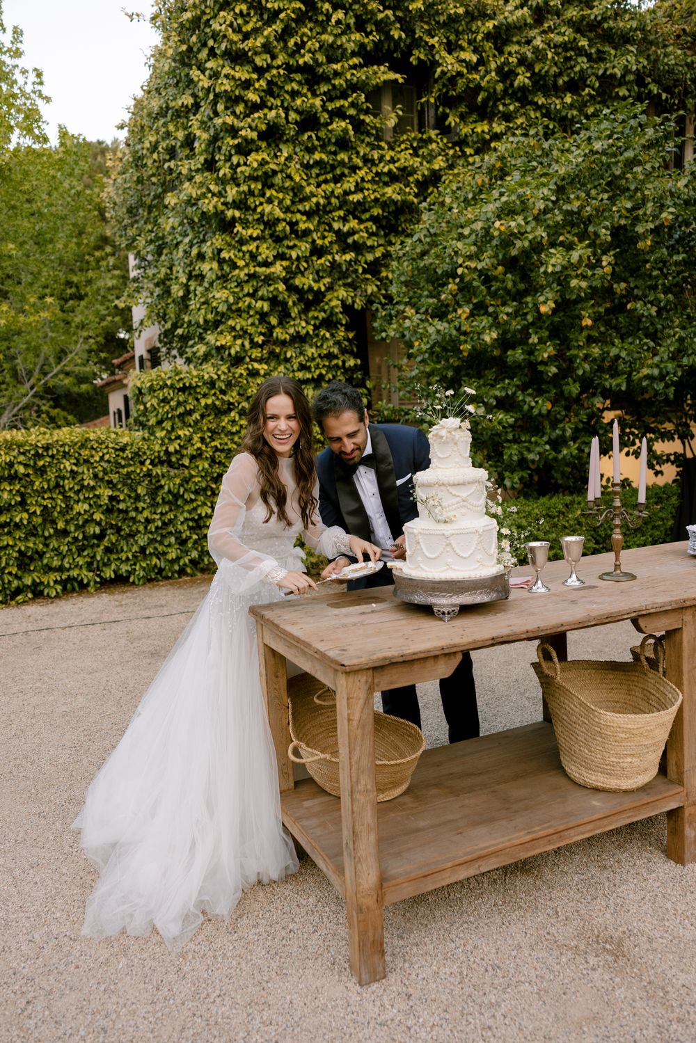 Newlyweds cut their elegant white tiered wedding cake together at a rustic outdoor table in a garden setting.