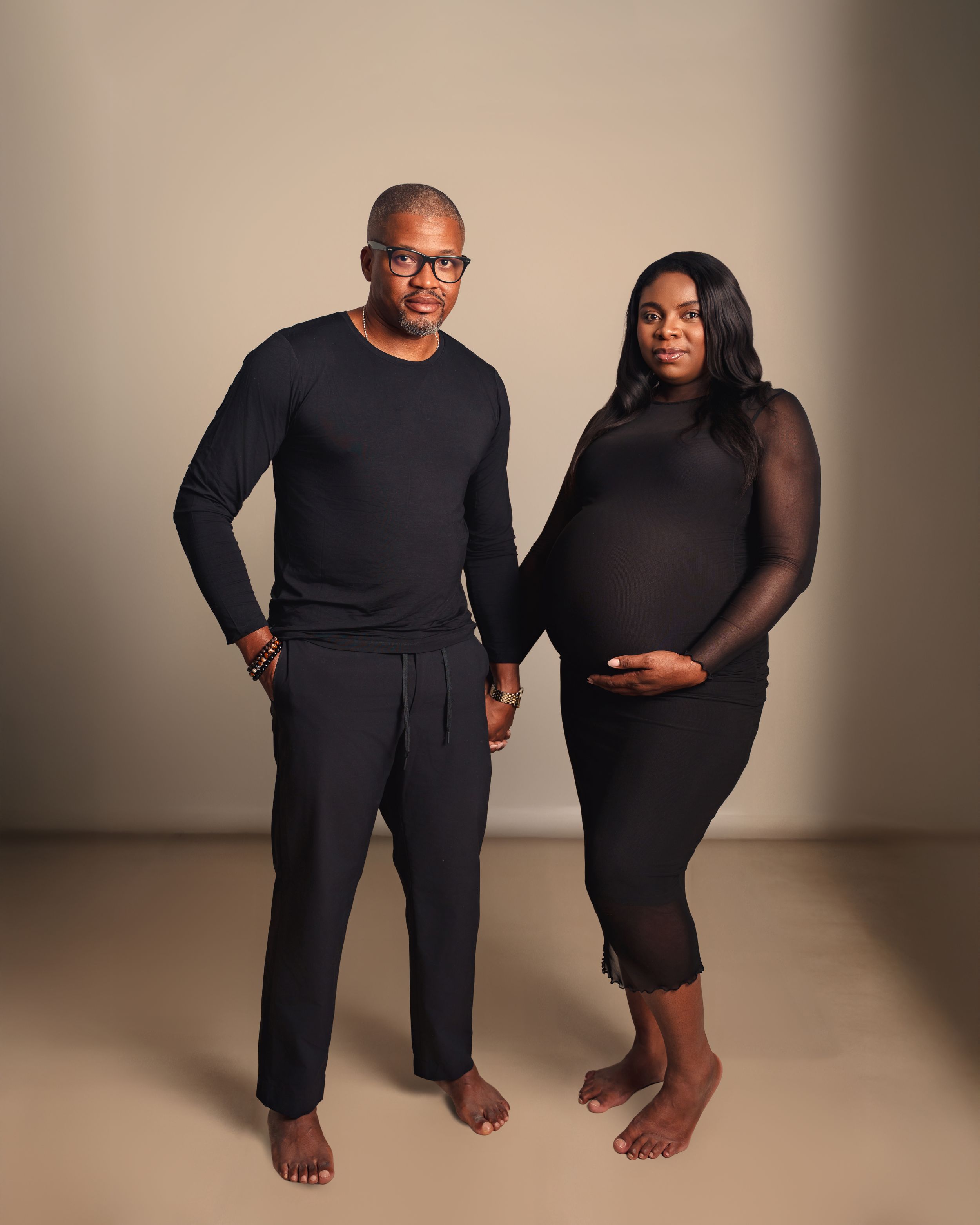 An expectant couple wearing black clothing poses together for a maternity photo against a neutral backdrop.