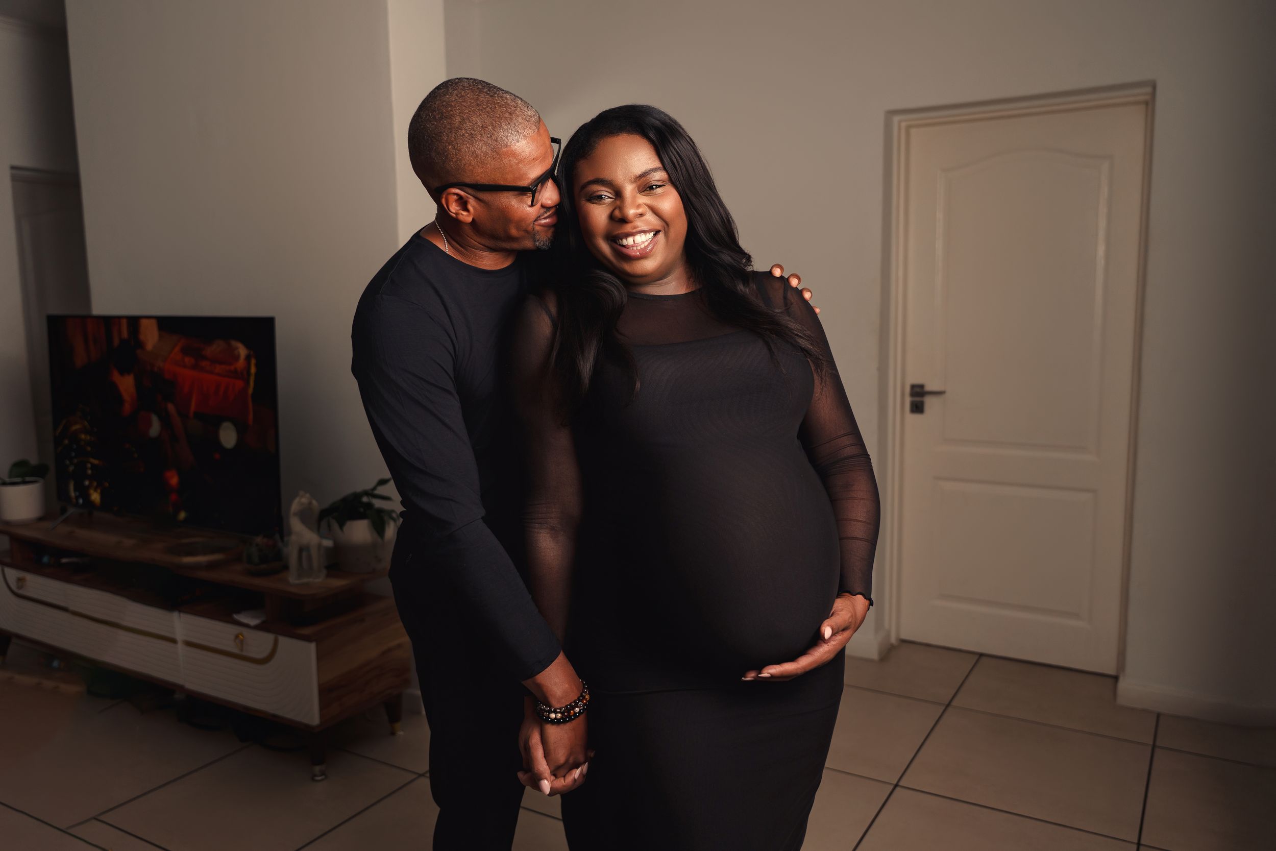 A couple wearing black outfits embraces warmly in an intimate photoshoot sequence against a white wall.