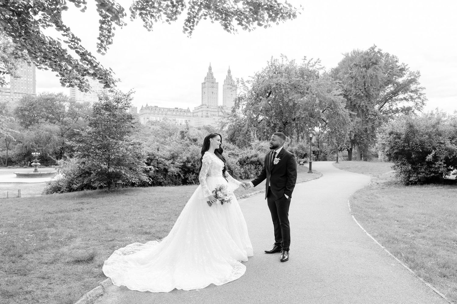 A couple in wedding attire poses on a winding park path with tall trees and historic architecture in the background.