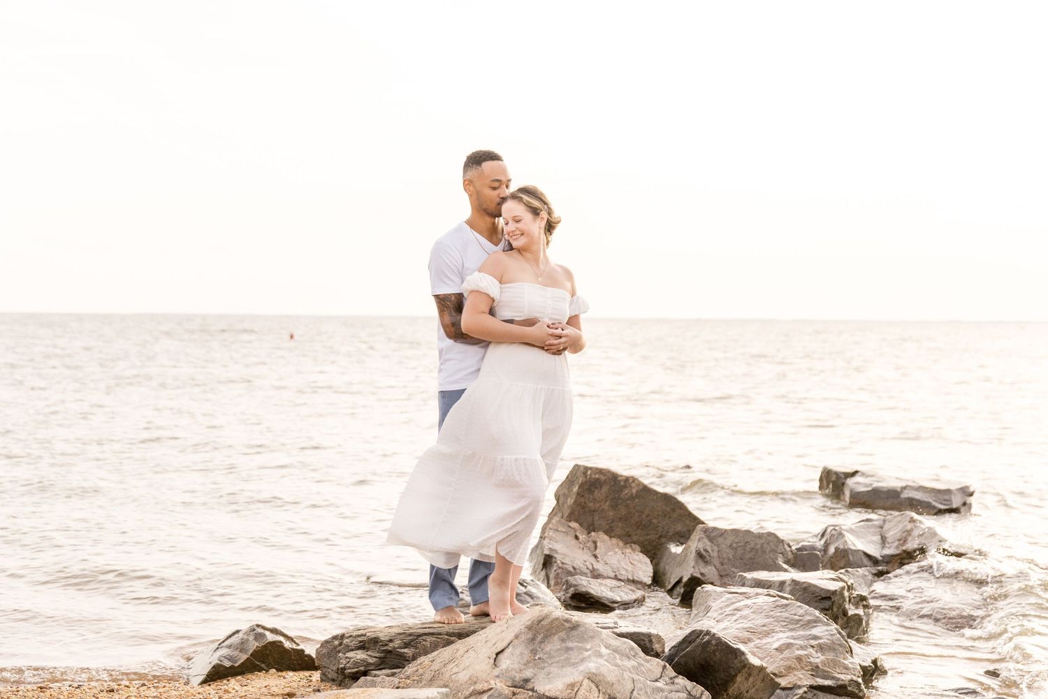 Couple in light wedding attire embrace on rocky beach at sunset, ocean waves splashing in background.