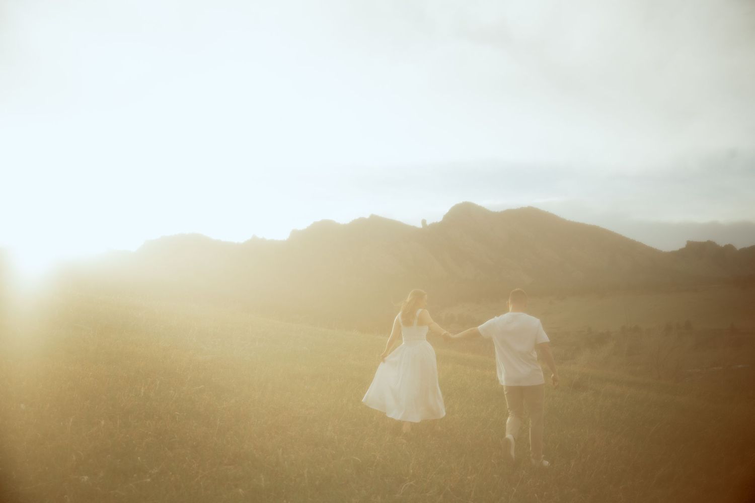 Two silhouetted figures run together through a mountain field at sunset with golden light glowing behind them.
