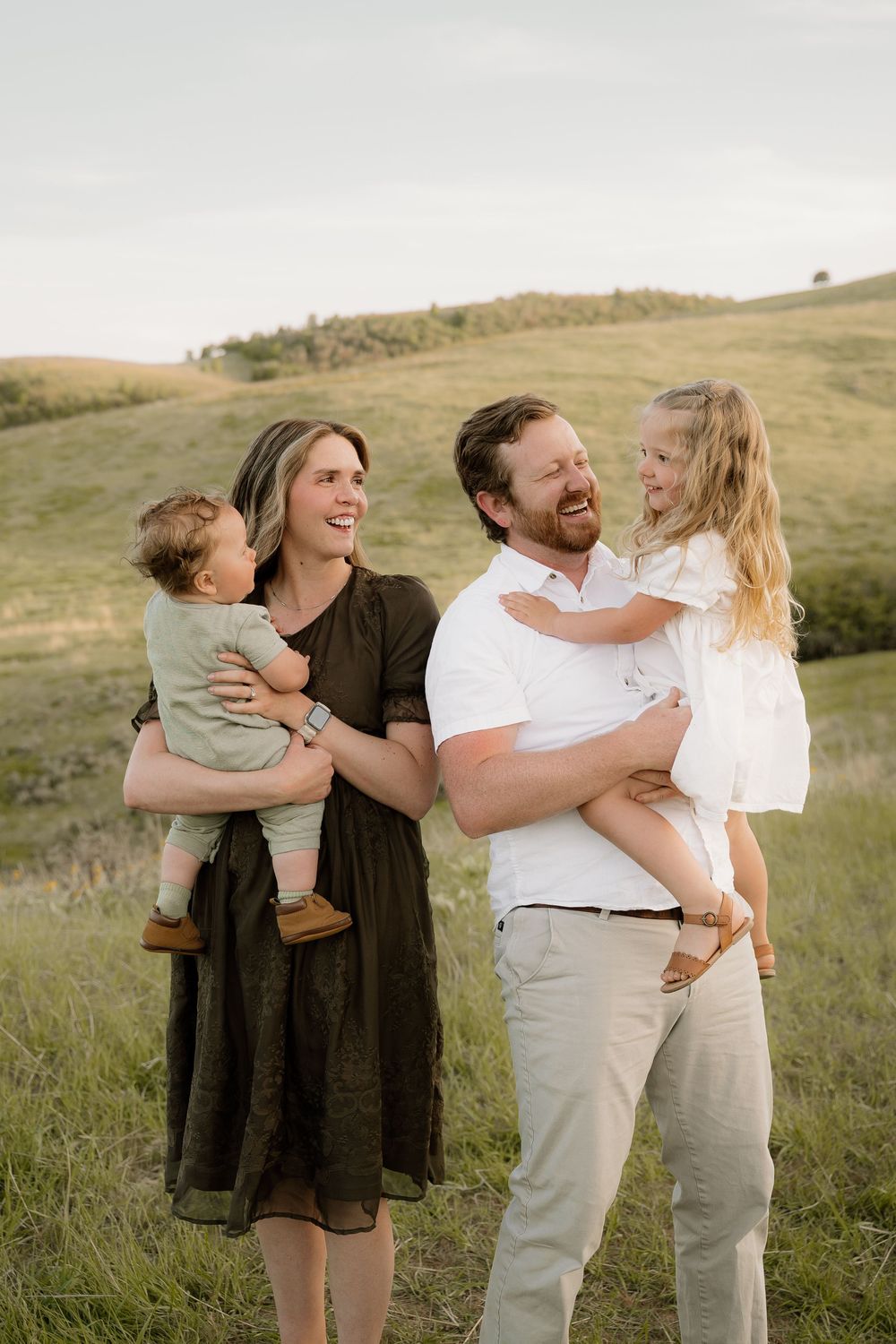 A family enjoys a candid moment together while standing in a grassy field during golden hour with rolling hills behind them.