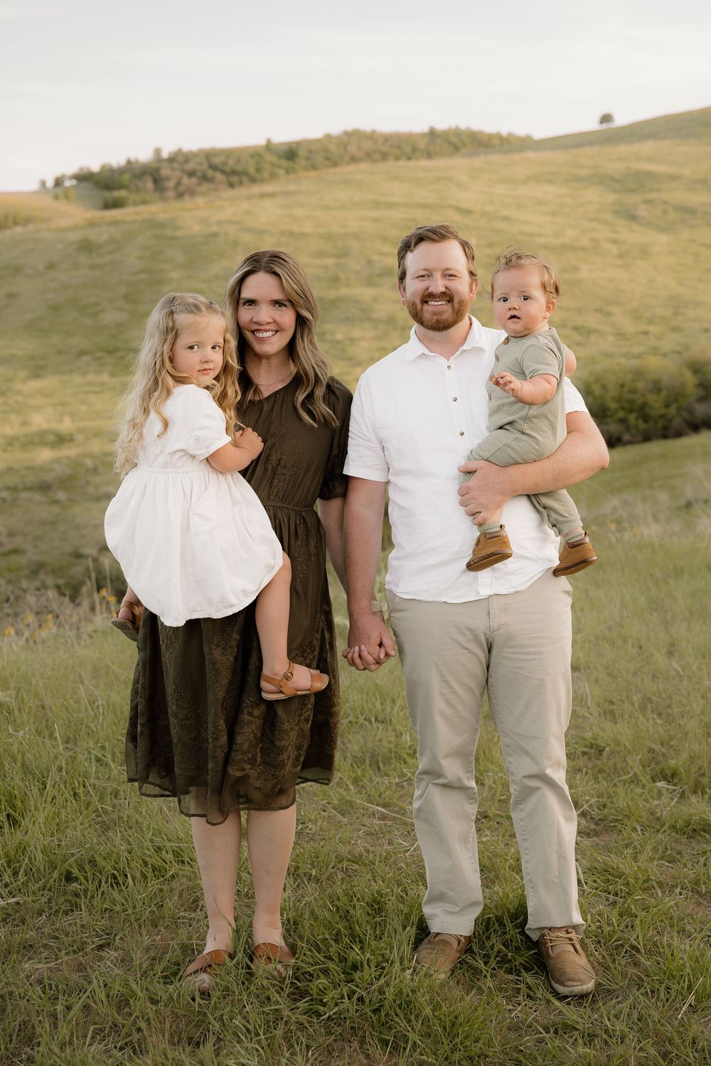 Family of four posing together outdoors on a grassy hillside during golden hour.