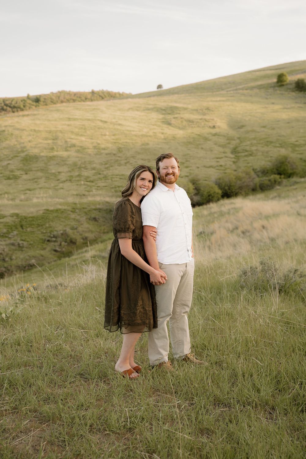 A couple stands together in a grassy hillside field on a sunny autumn day wearing earth-toned clothing.