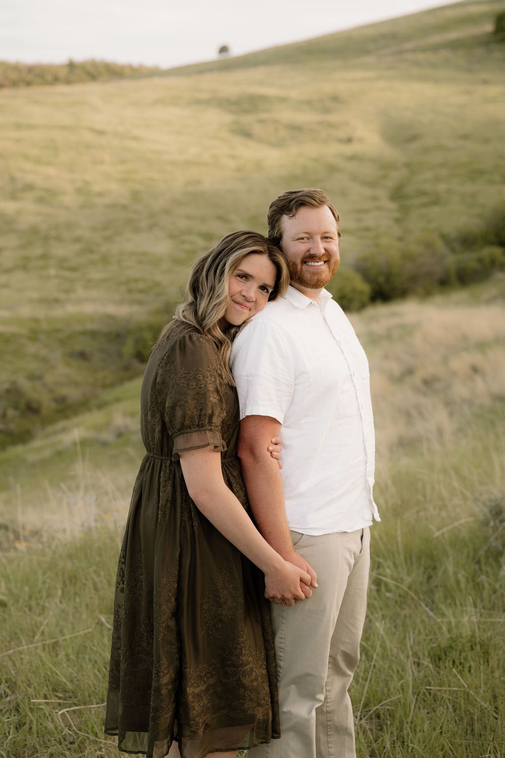 A couple stands together in a grassy field wearing white and brown outfits at sunset.