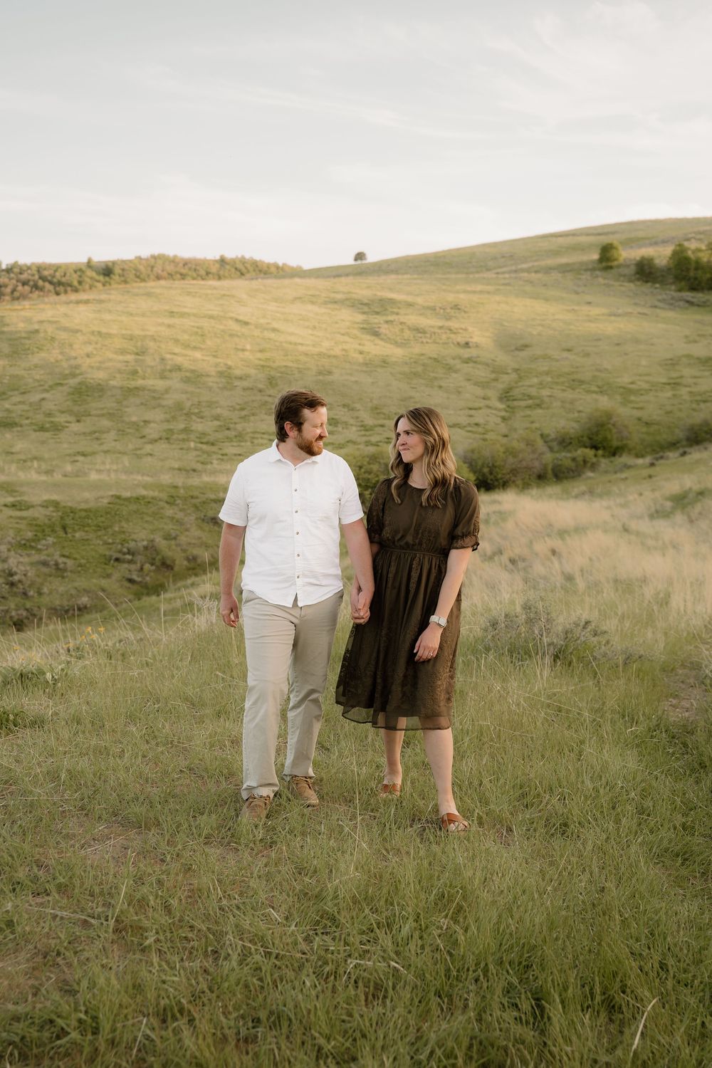 Two people stand together in a grassy hillside field at sunset wearing casual attire.