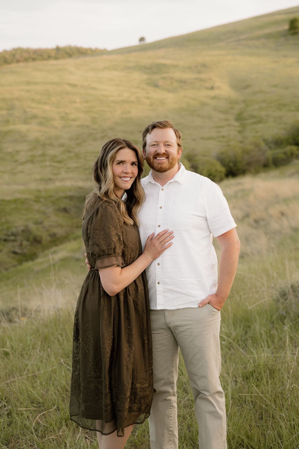 A couple poses together on a hillside wearing coordinated earth-toned outfits at sunset.