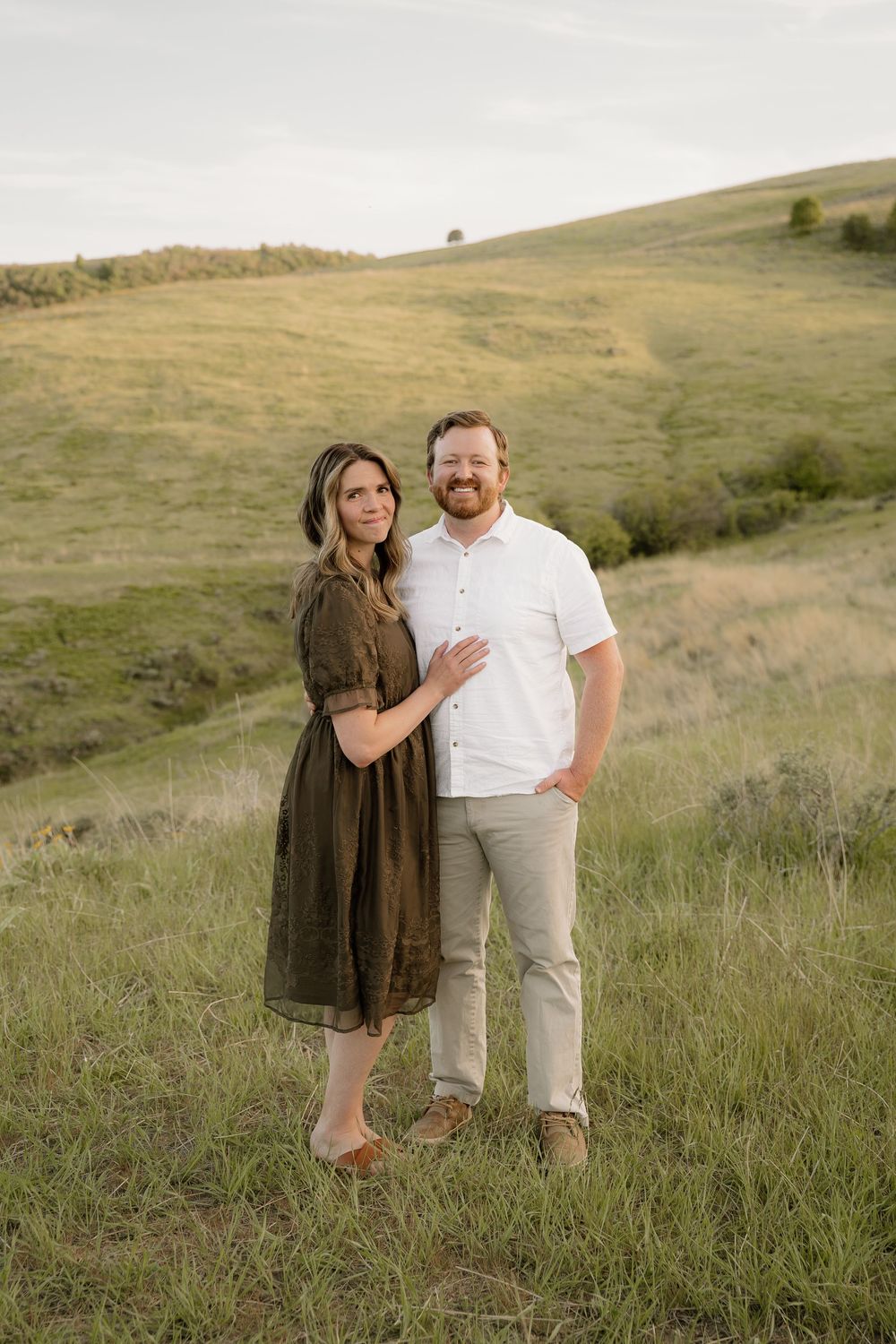 Couple poses affectionately in golden grassy hillside wearing white and olive brown outfits during sunset photoshoot.
