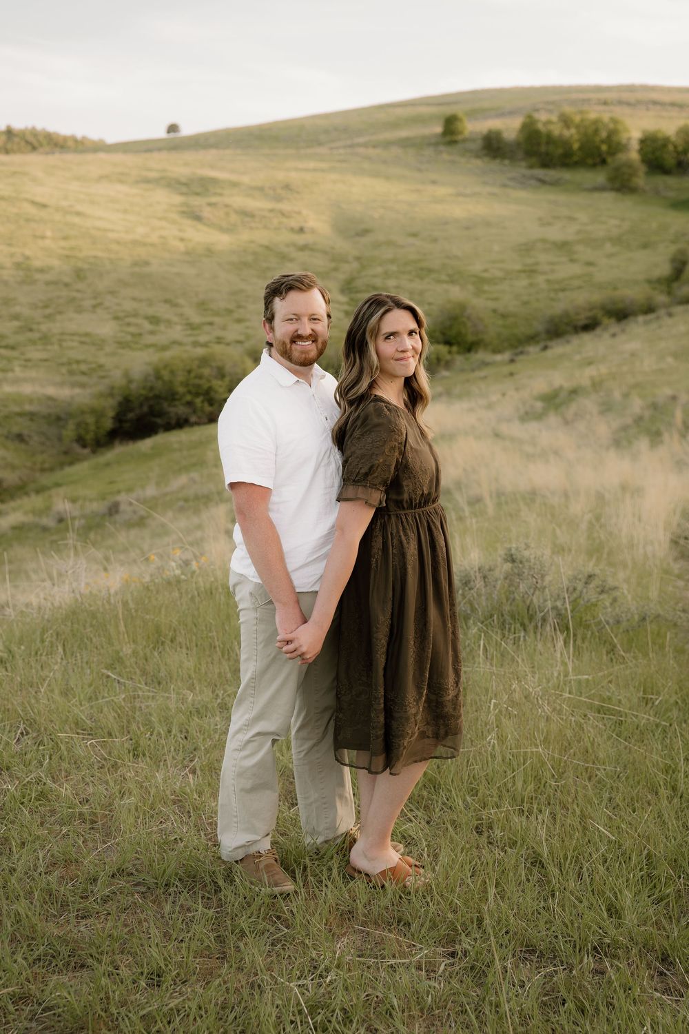 Two people stand close together in a serene hillside meadow wearing coordinated white and brown casual attire.