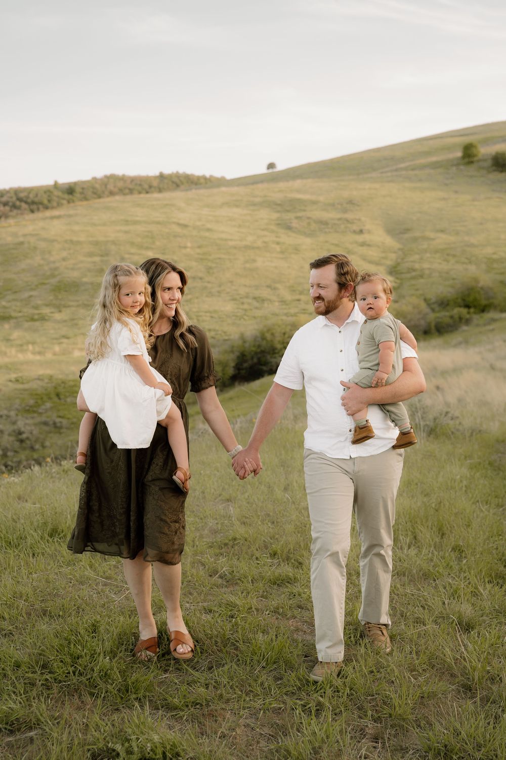 Family walks together holding hands across scenic grassy hillside during golden hour photo session.