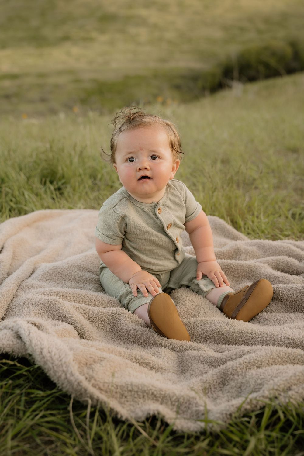 A baby in a tan outfit sits on a blanket in tall grass on a hillside.