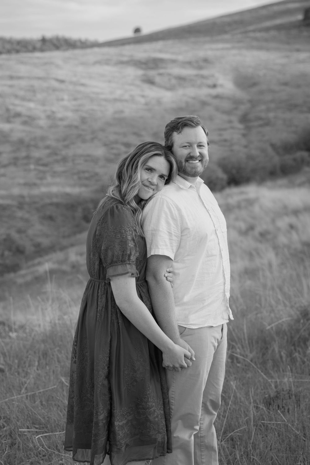 Black and white outdoor portrait of a couple embracing on a hillside with tall grass in a natural setting.