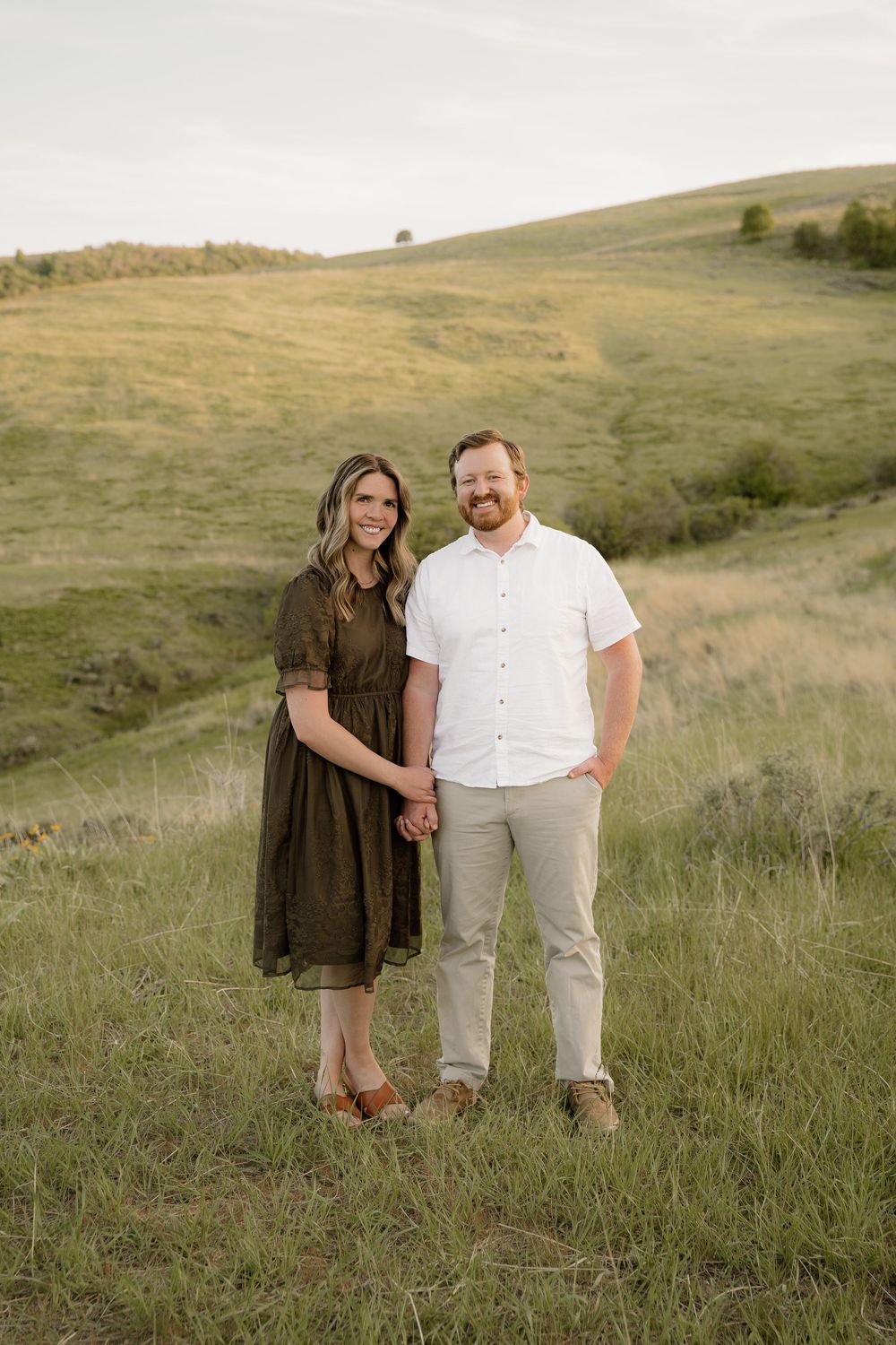 A couple stands together on a grassy hillside during golden hour, wearing casual dressy outfits in earth tones.