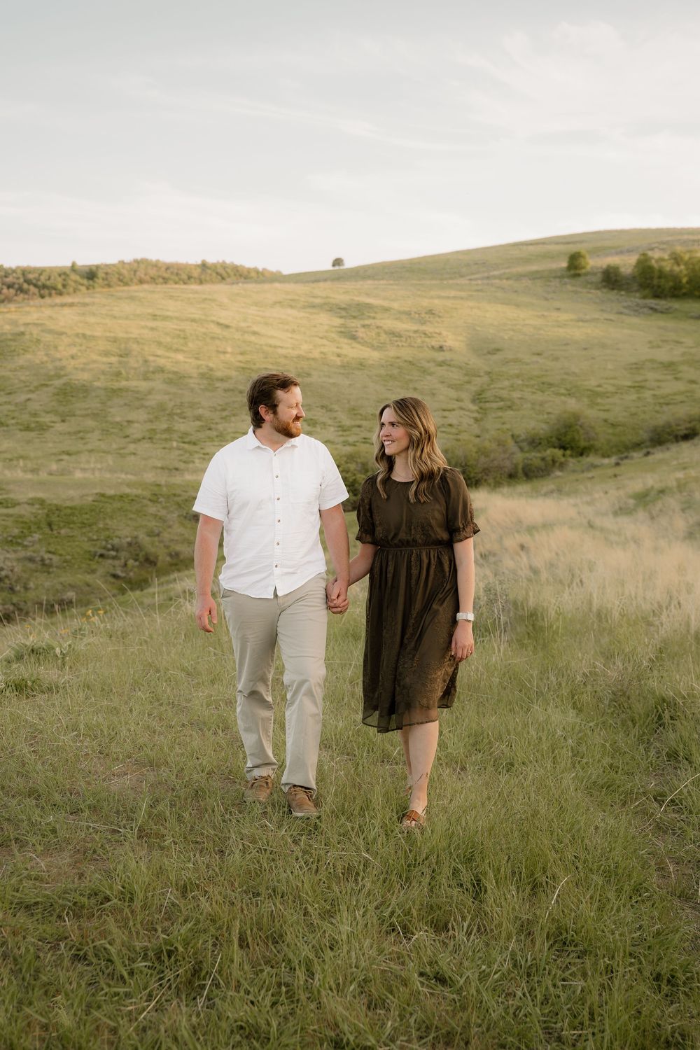 A couple walks hand in hand through tall grass on a scenic hillside at sunset.