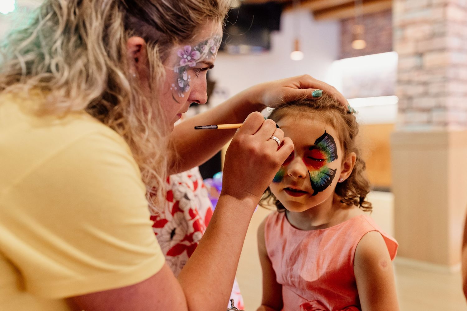 Artist painting colorful face paint designs on young child at a party with vibrant colors and festive atmosphere.