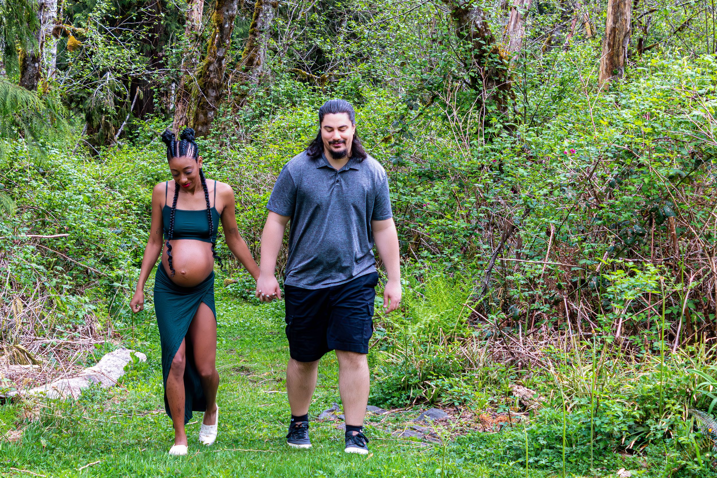 A pregnant woman holds hands with a man as they walk through the forest.