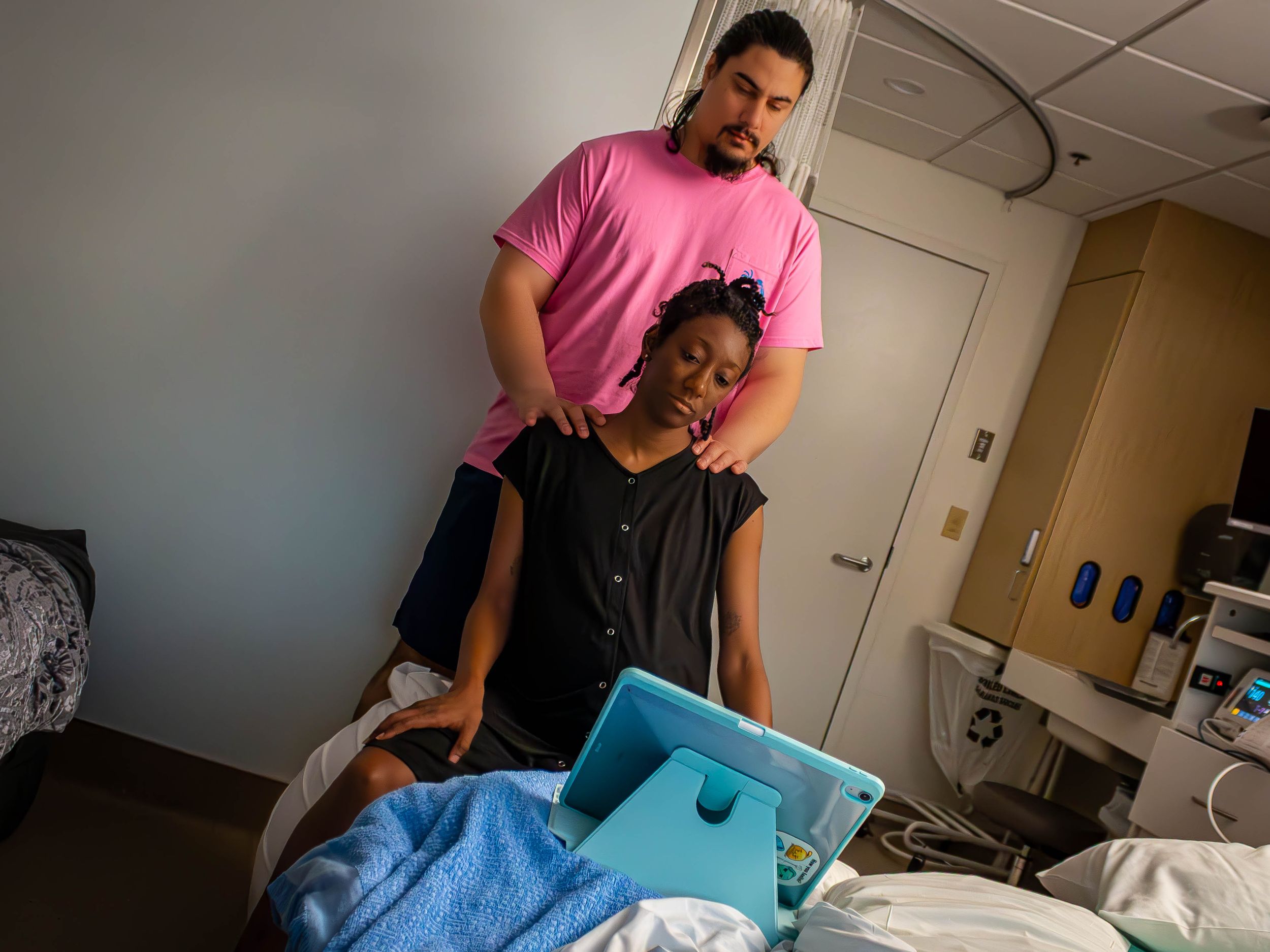 Two people in a hospital room, with one person standing and another sitting on a medical bed with blue sheets.