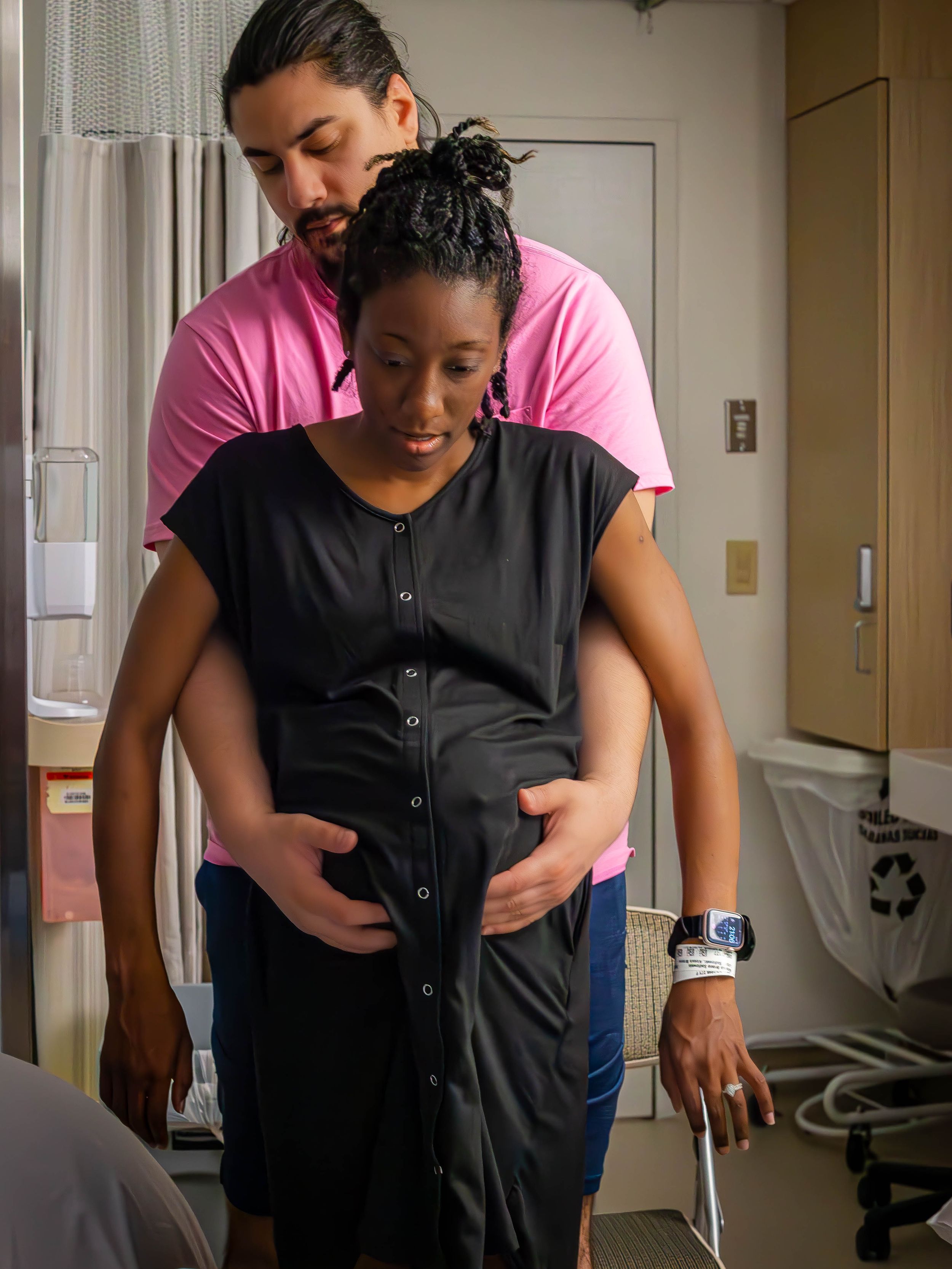 Dad-to-be stands behind mom-to-be and lifts her belly in a hospital room during active labor.