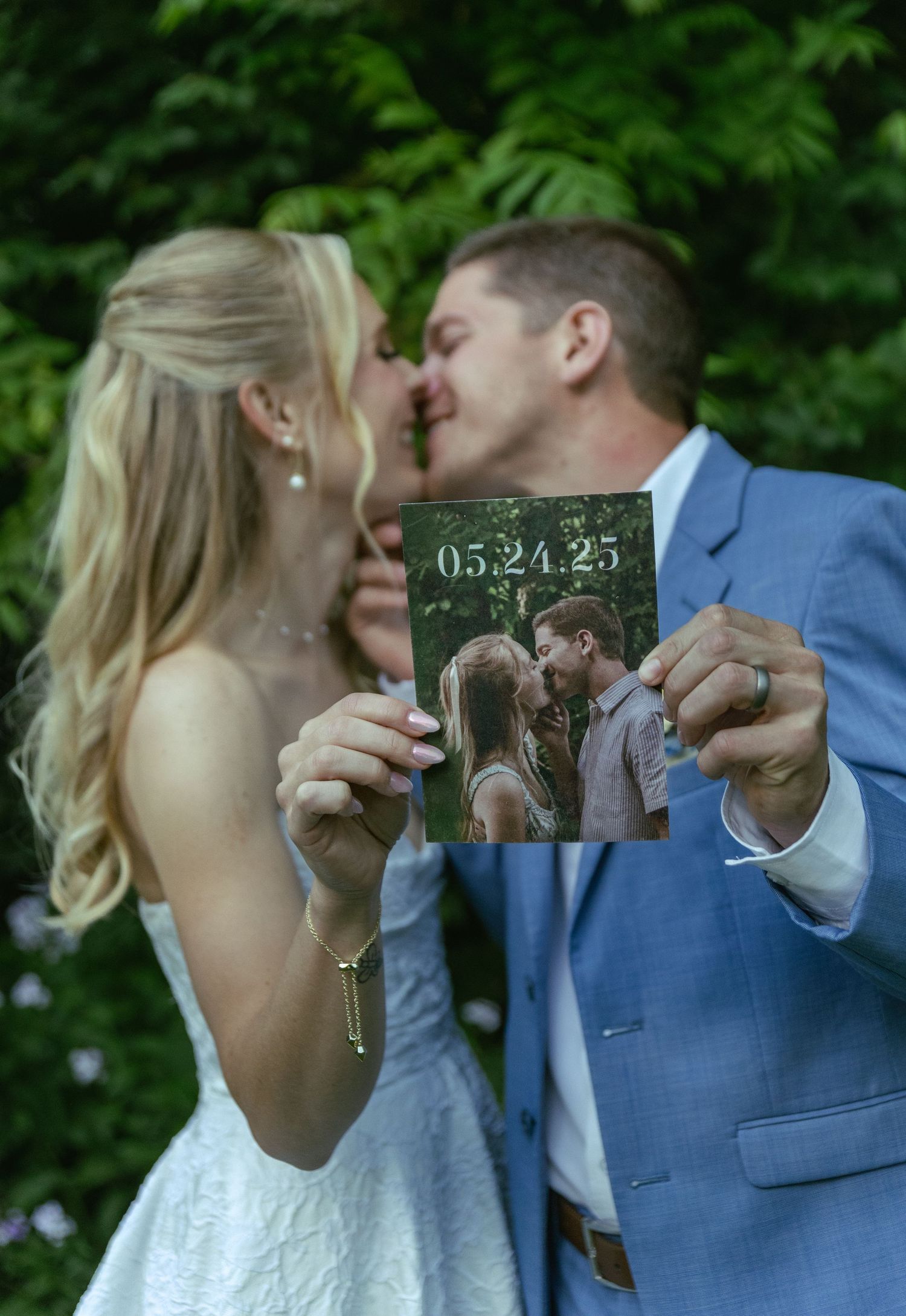Newlyweds share a kiss while holding their wedding date sign outdoors.