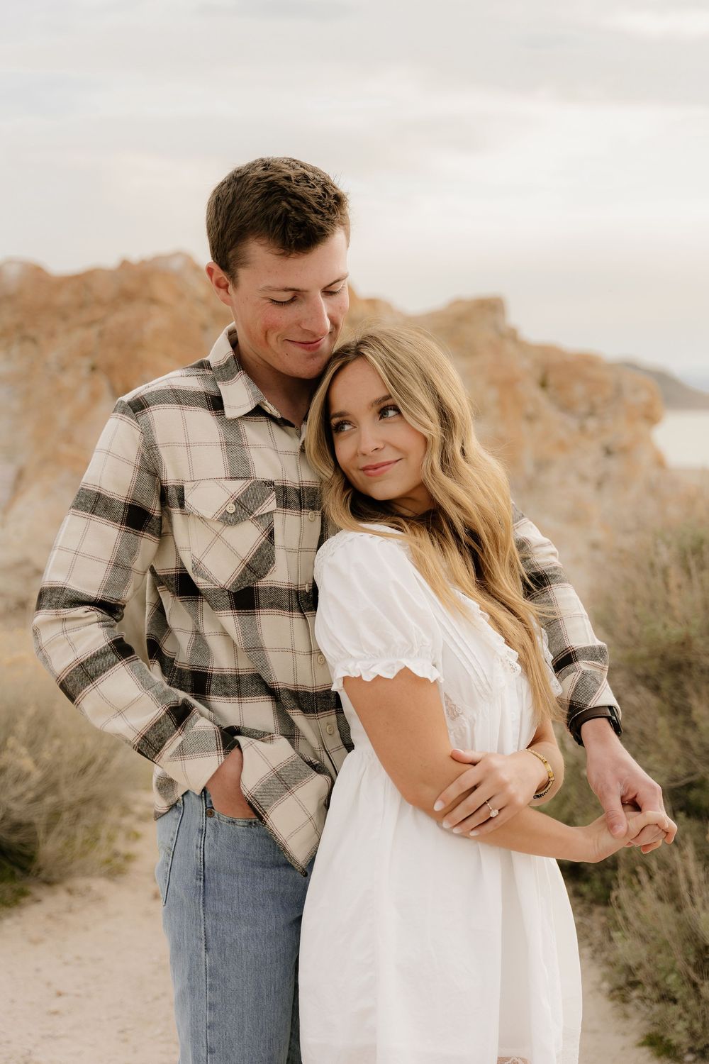 A couple embraces on a desert trail with mountain cliffs in the background during golden hour.