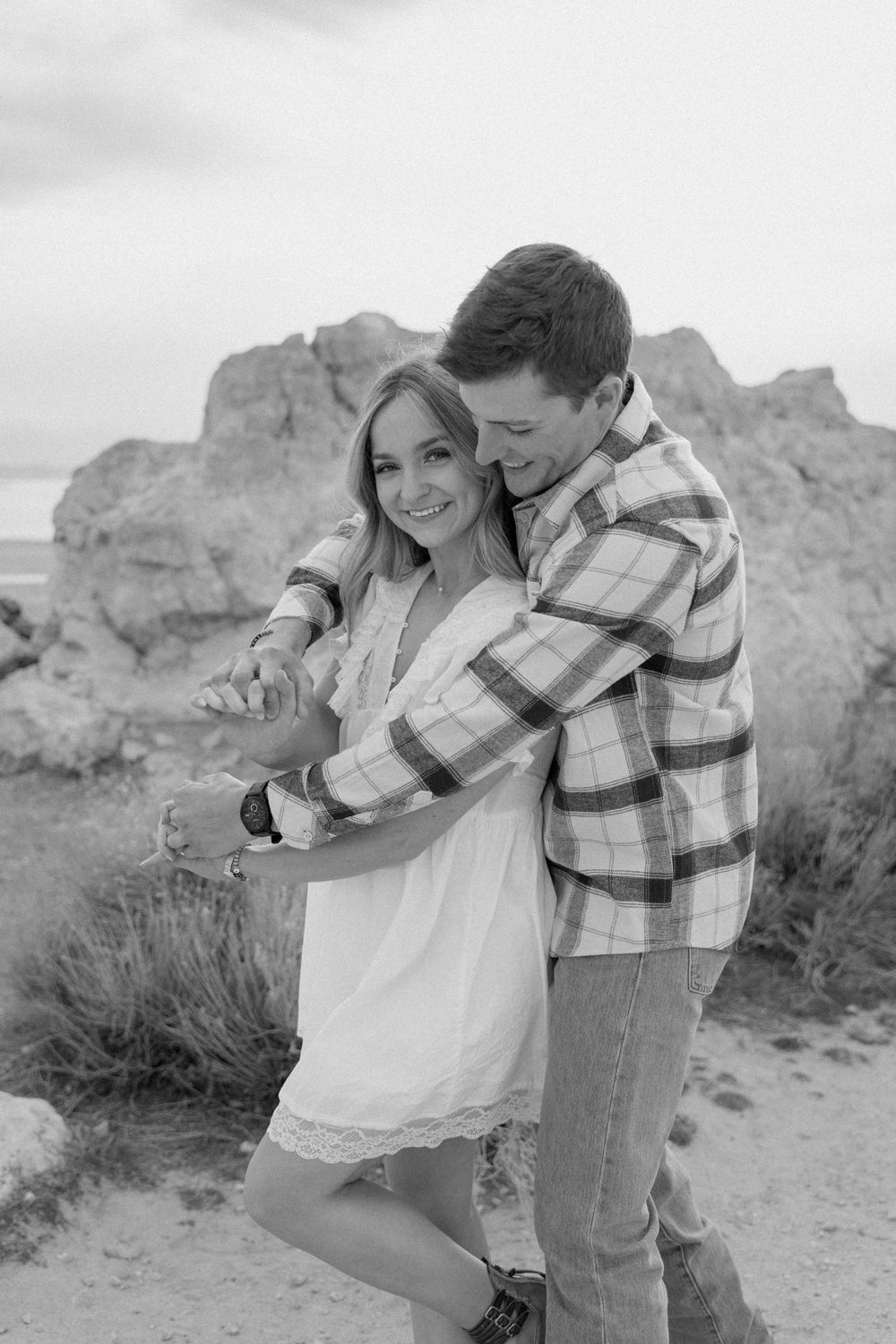 A couple embraces while posing together in a desert landscape with rocky formations in the background.