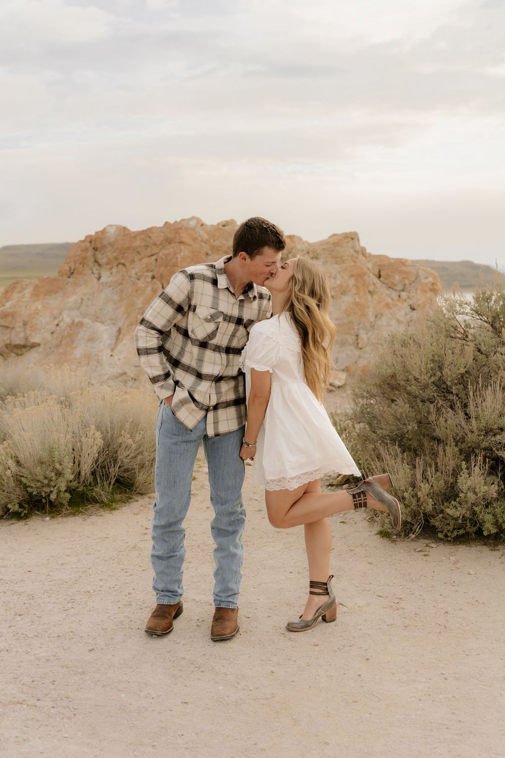 A romantic couple shares a tender moment on a dirt path with rugged desert rock formations and brush in the background.