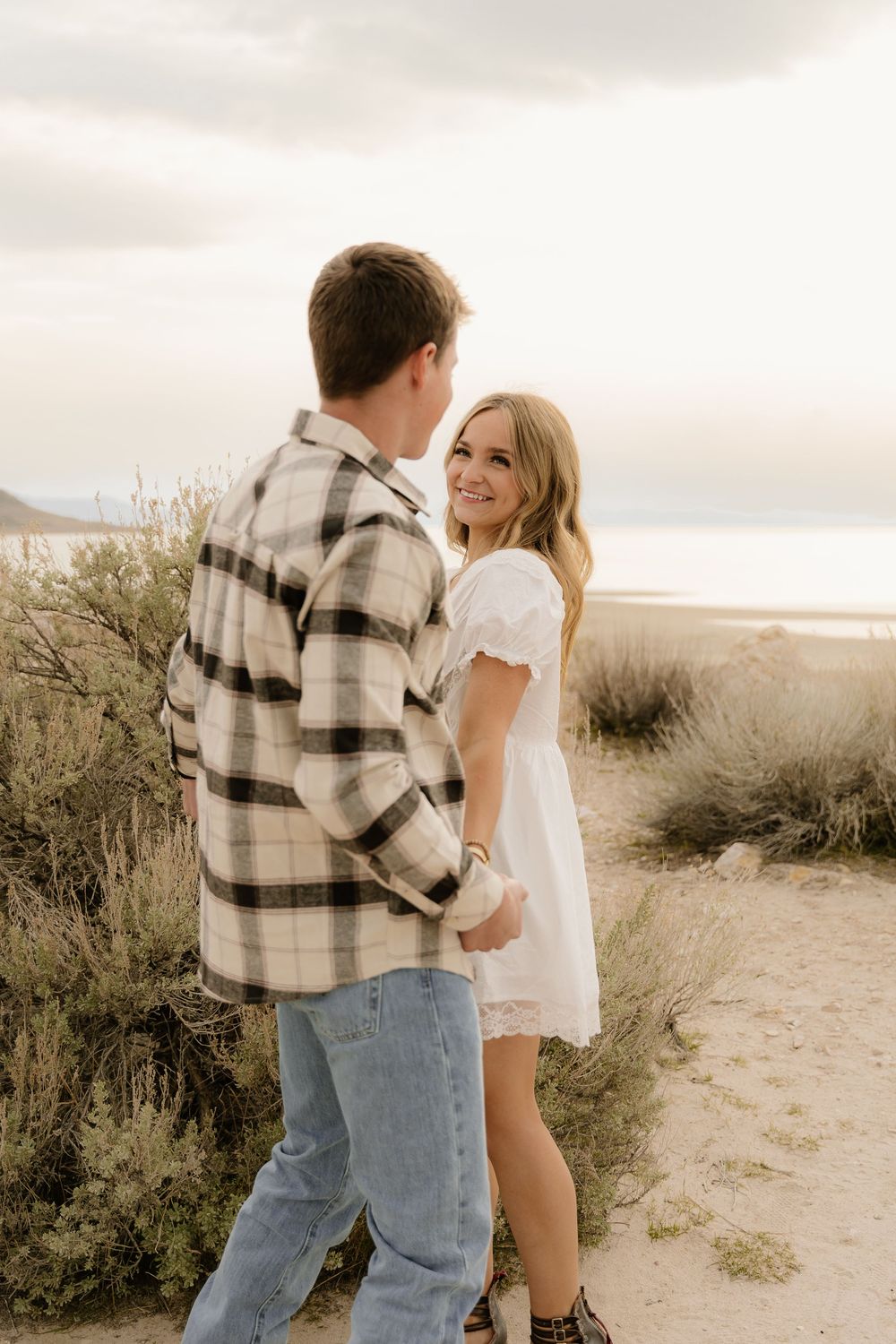 Couple shares intimate moment on desert trail with mountains and brush in background at sunset.