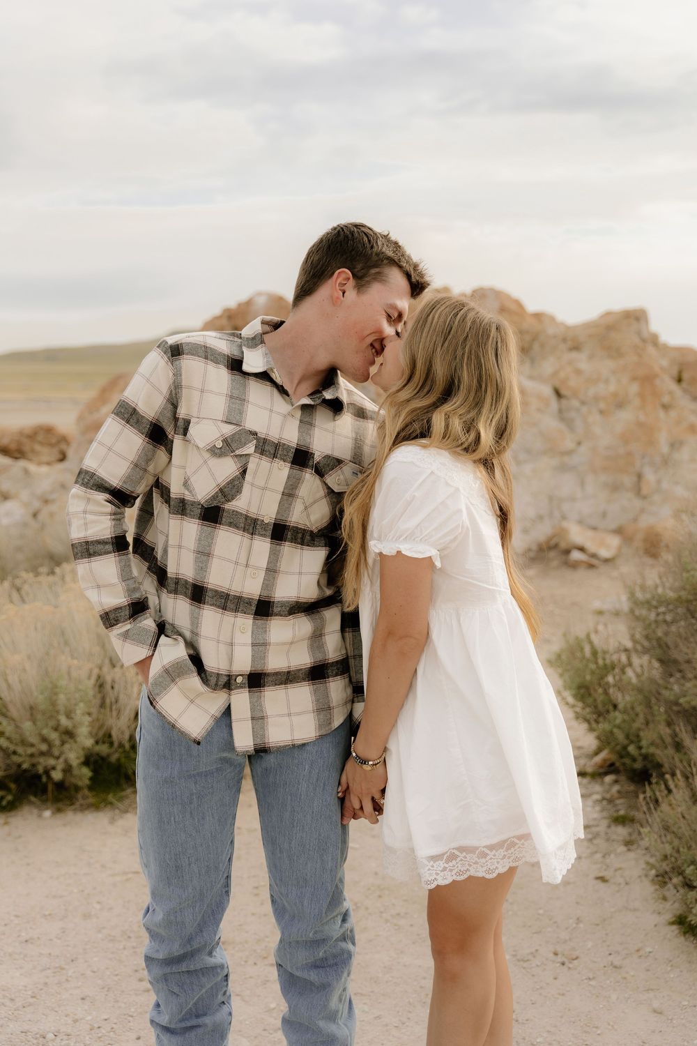 A couple in casual western attire share an intimate moment on a desert trail with rocky formations in the background.