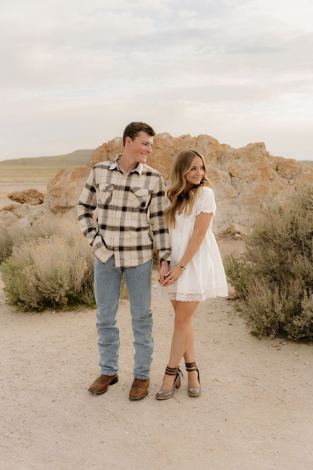 Two people hold hands in an intimate outdoor desert setting with rocky formations and brush in the background at sunset.