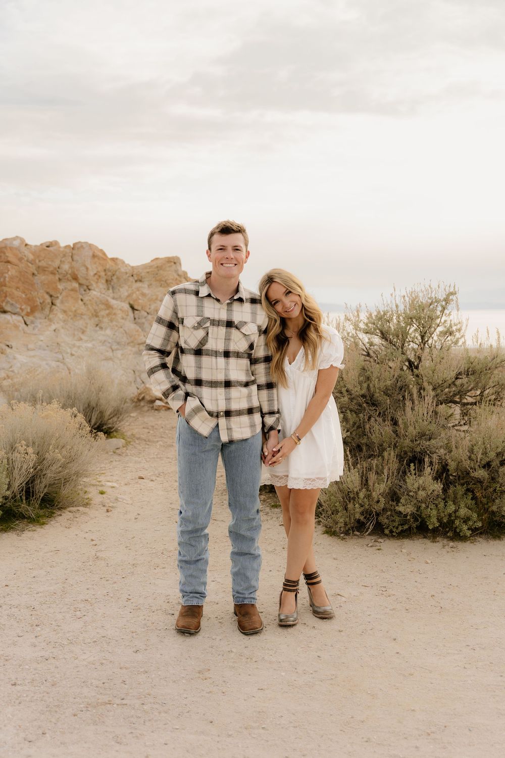 A couple poses together on a desert trail at sunset wearing casual western-inspired outfits.