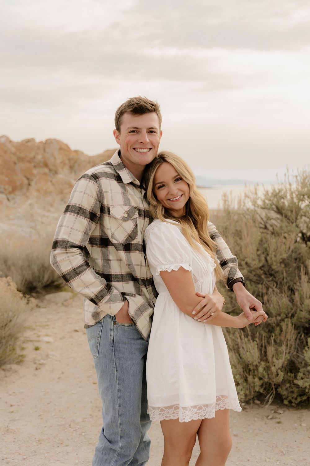 A couple poses together on a desert trail with rocky mountains and sage brush in the background.