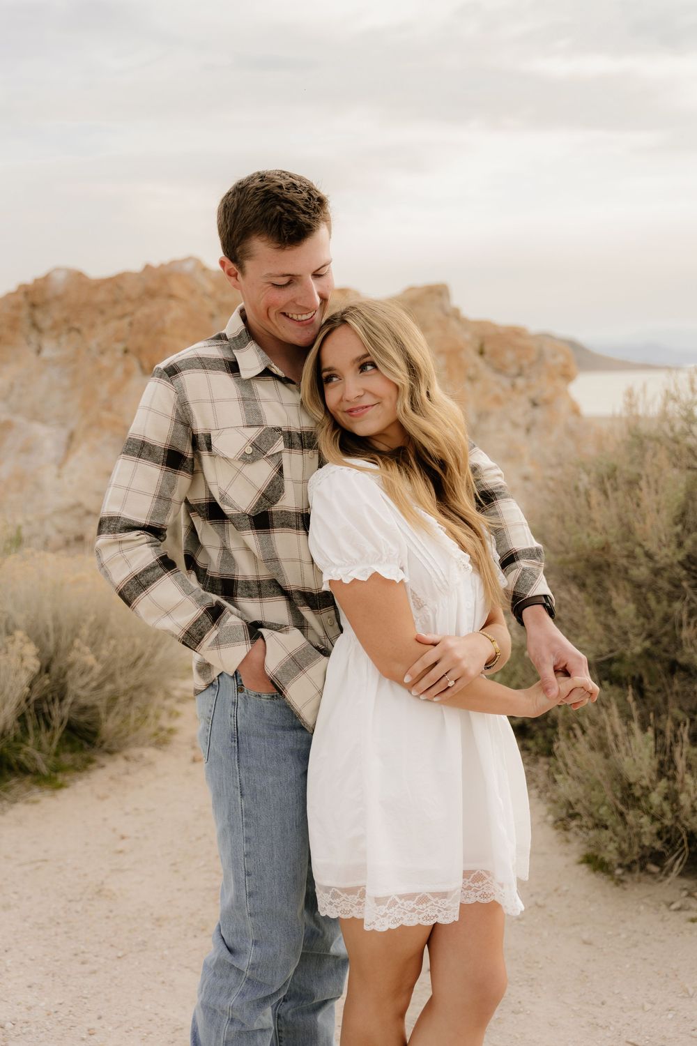 A couple shares an intimate moment in a desert landscape wearing a plaid shirt and white dress.