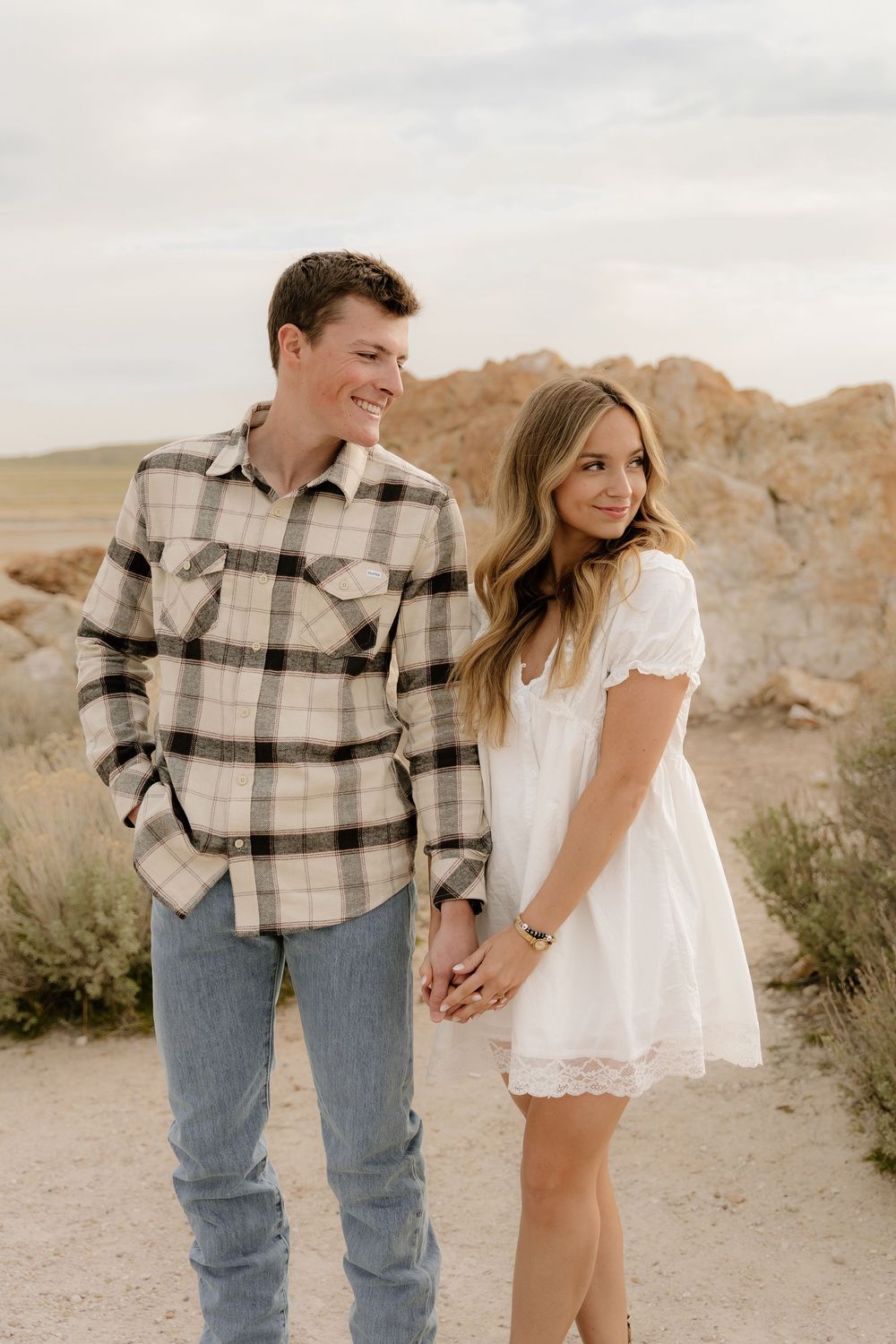 A couple stands together on a desert trail, with the person in plaid holding hands with someone in a white dress.