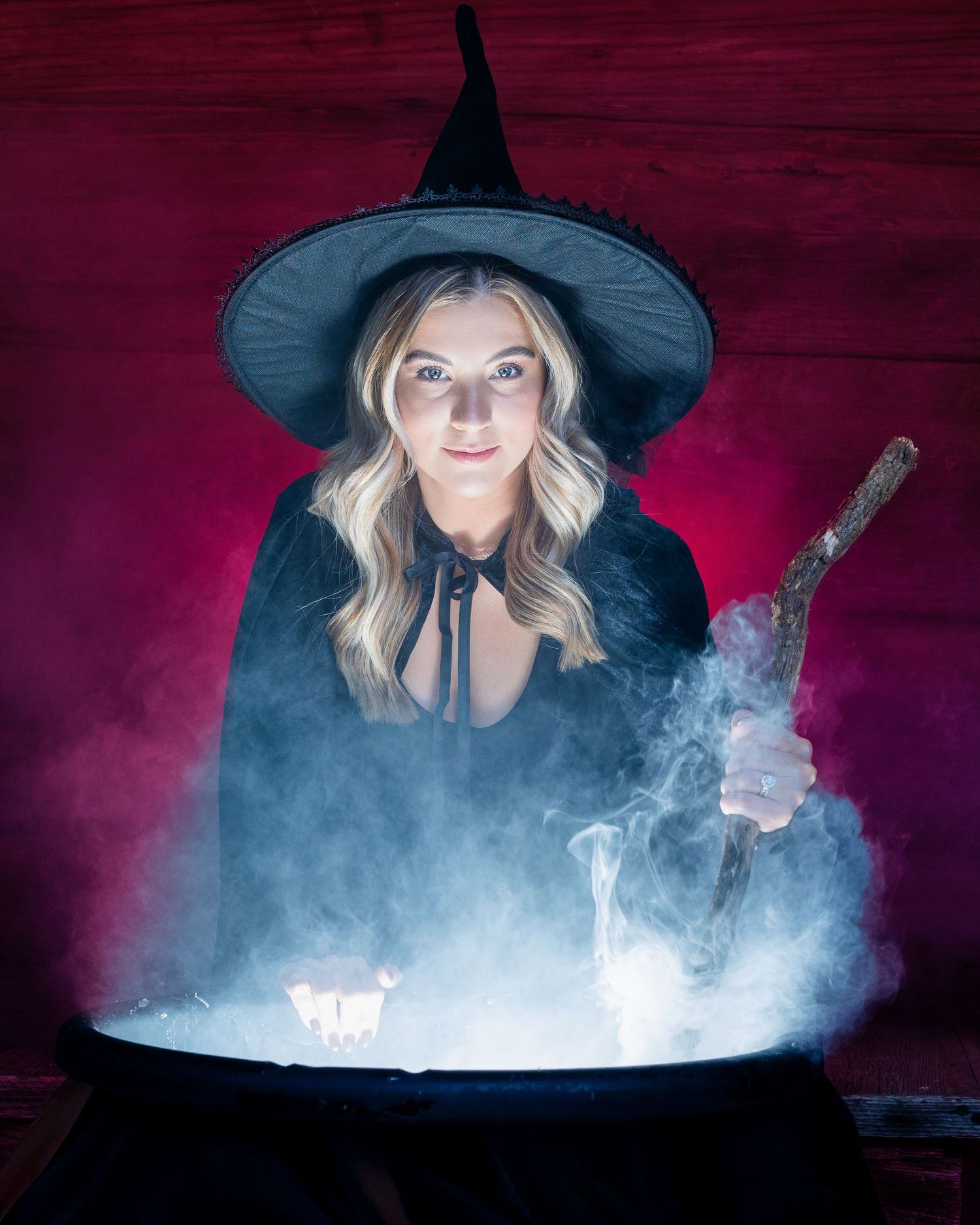 Figure in witch hat and black cloak stirring misty cauldron against dark red background.