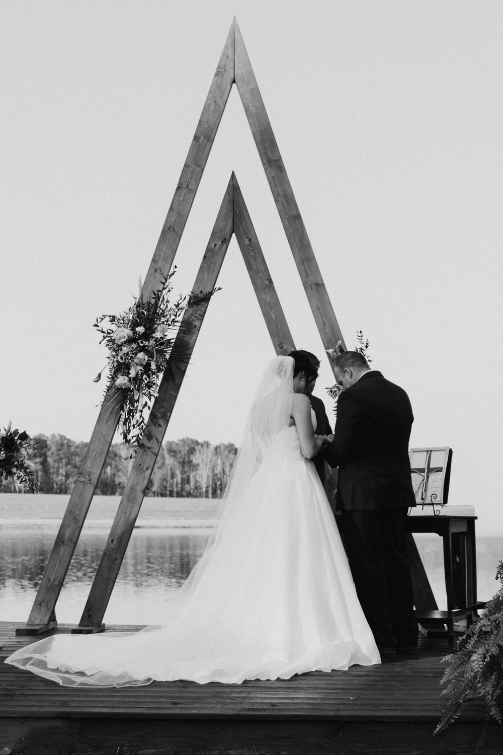 Black and white wedding photo under a geometric arch by a serene lake with couple tying the symbolic knot.