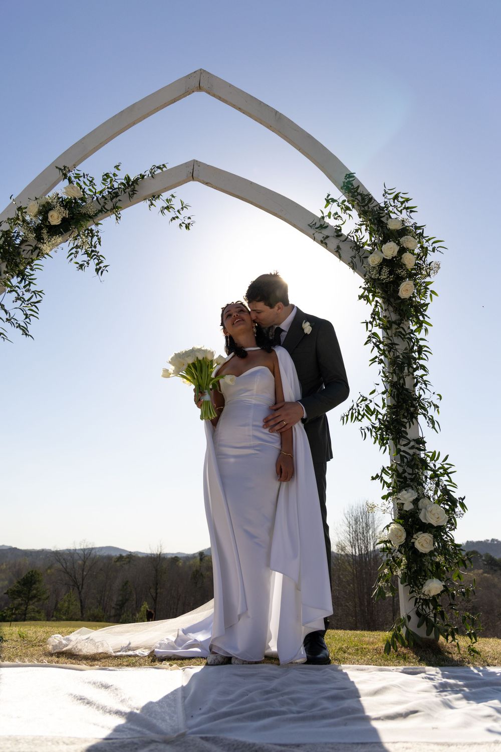 Wedding ceremony under a floral arch with mountain backdrop and sunlight streaming through.